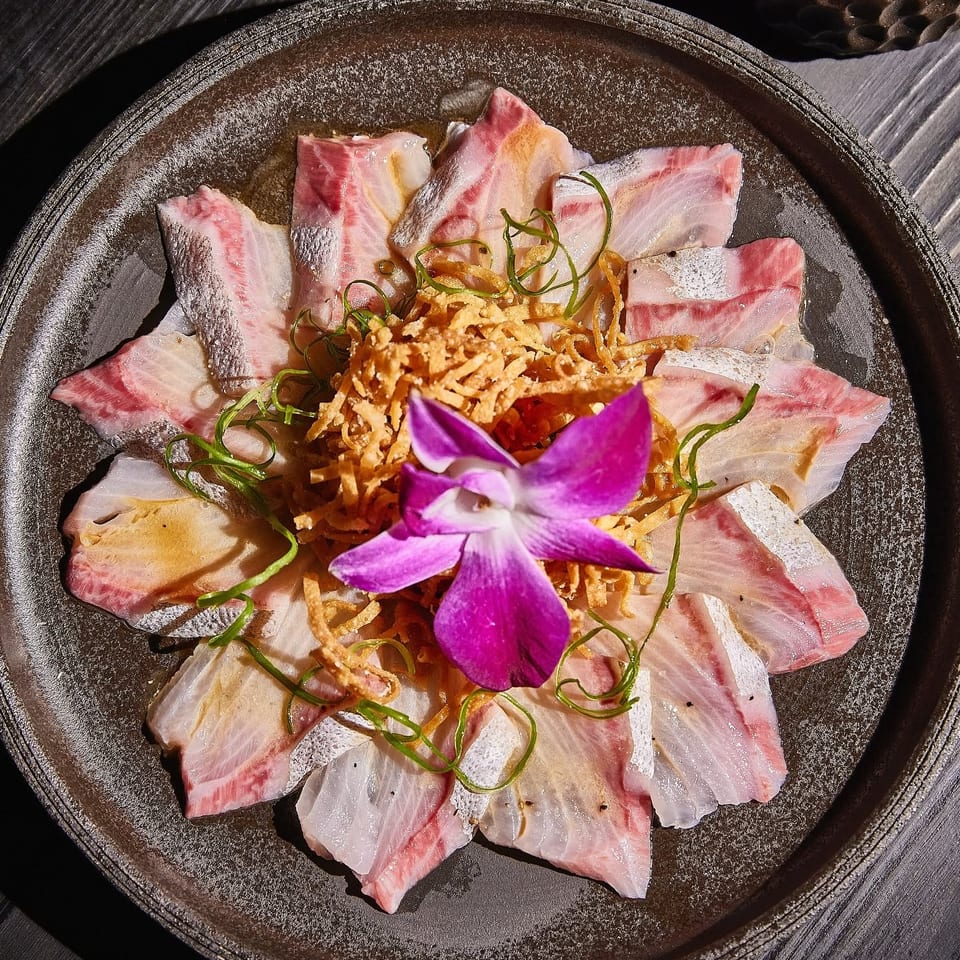 Thinly sliced pink fish arranged around crispy garnish, scallions, and a purple orchid on a dark ceramic plate.