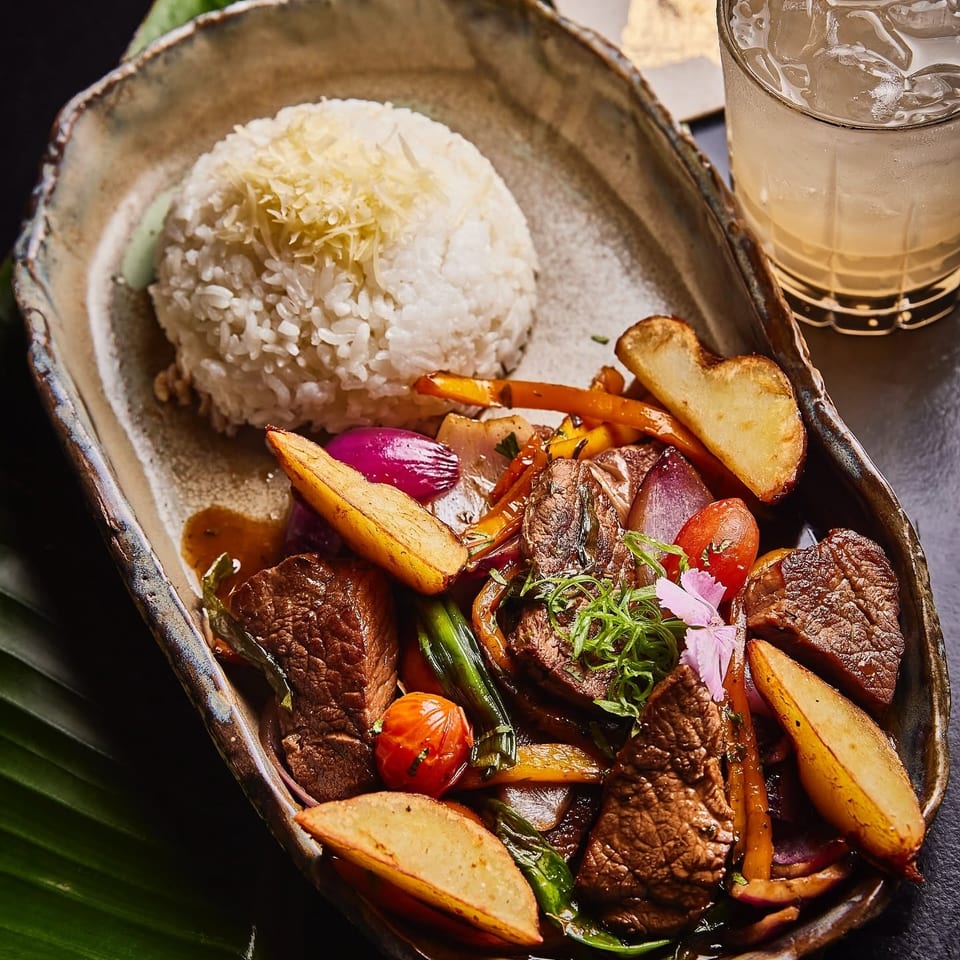 Seared beef with roasted vegetables and potato wedges served beside jasmine rice, a citrus-garnished iced drink, and tropical leaves.