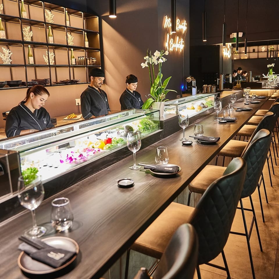 Modern Japanese restaurant interior with chefs behind a sushi counter, fresh ingredients, floral arrangements, and neatly set bar seating.