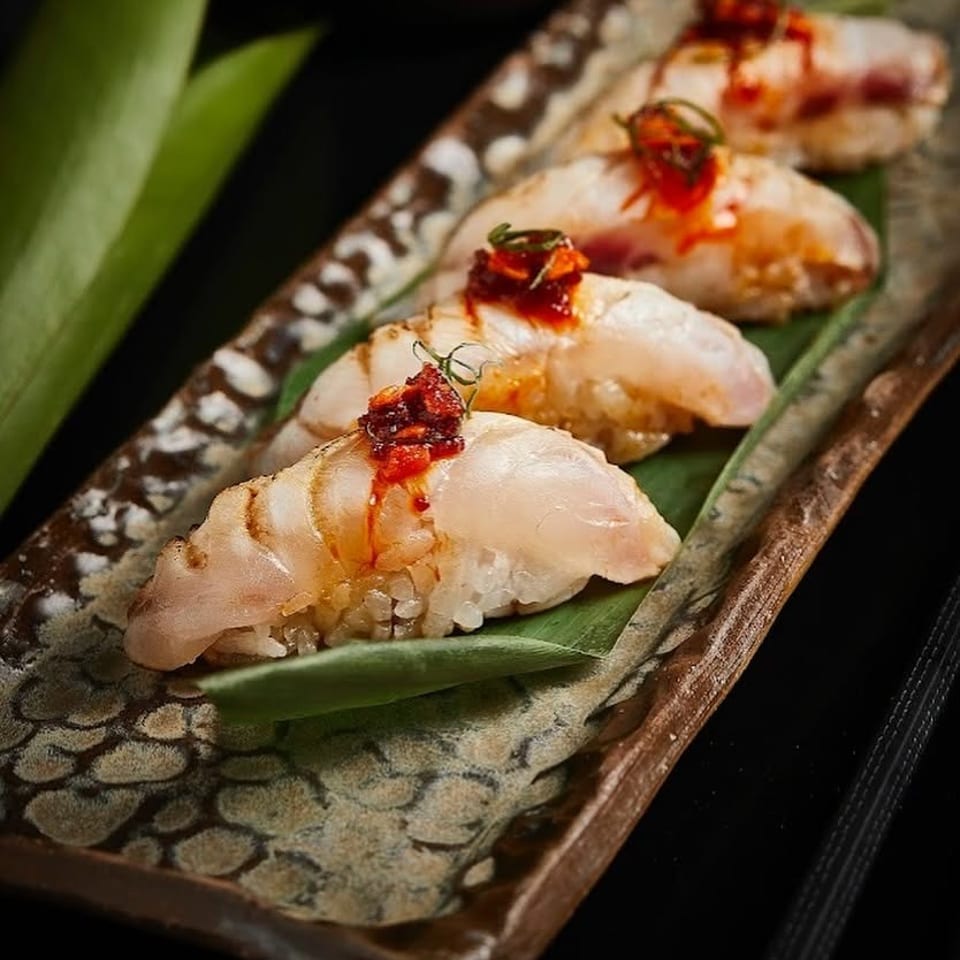 Four pieces of glazed fish nigiri topped with red chili relish and herbs on a leaf-lined ceramic platter.