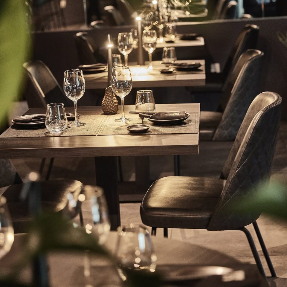 Warmly lit restaurant dining room with elegantly set wooden tables, wine glasses, candles, and upholstered chairs.