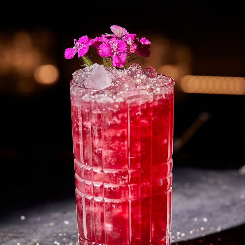 Vibrant red cocktail in a textured glass, topped with crushed ice and bright pink flowers against a dimly lit bar background.