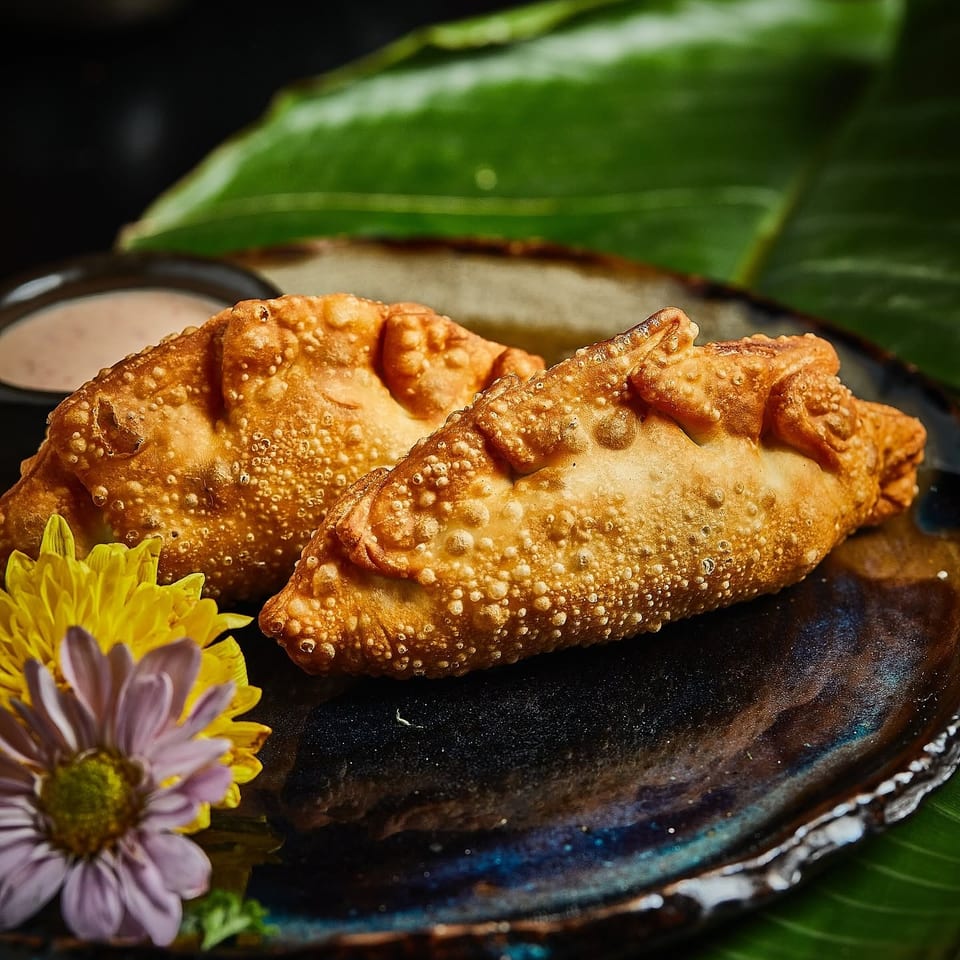 Golden, crispy folded pastries served on a dark plate with dipping sauce, green leaves, and flowers.