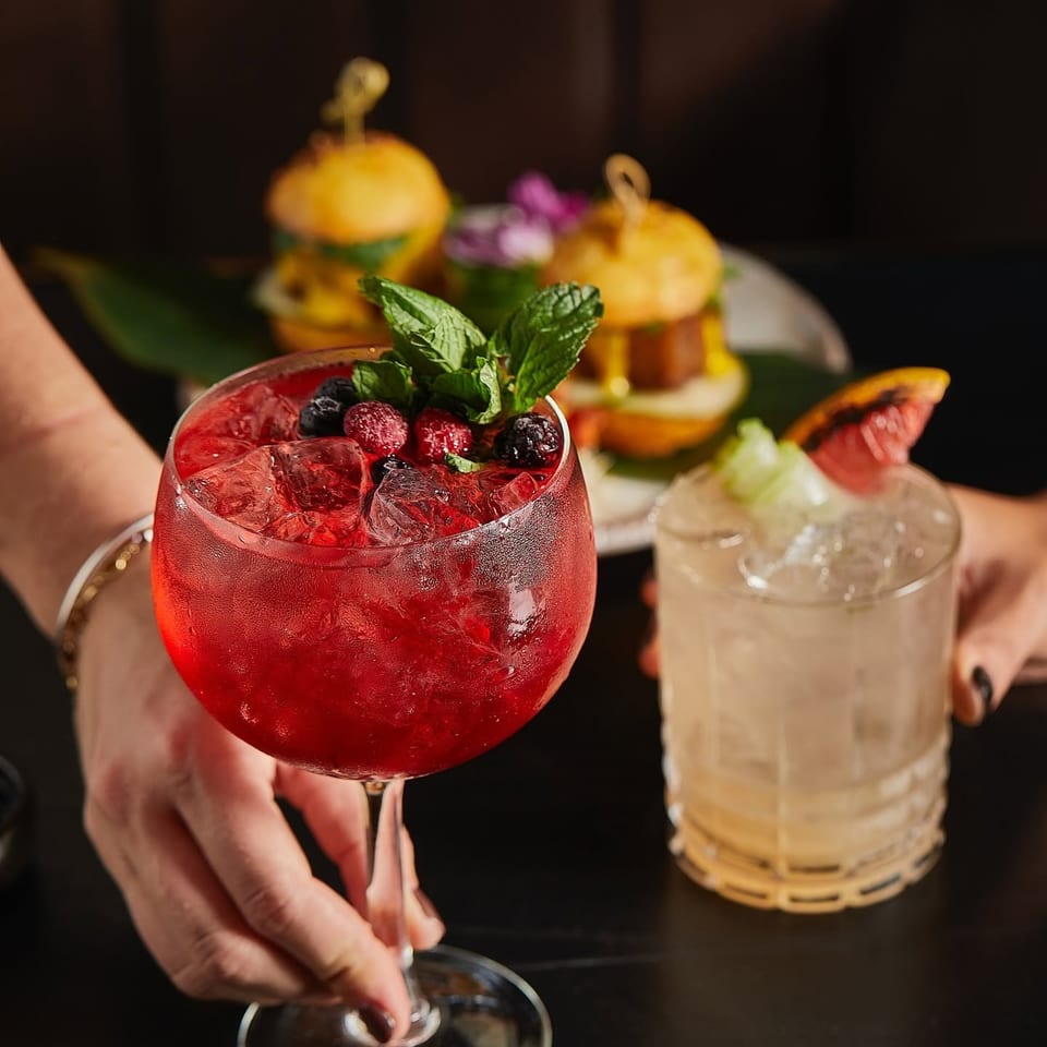 Hands hold a berry-filled red cocktail garnished with mint, alongside a pale drink and sliders on a restaurant table.