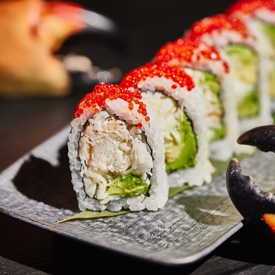 Close-up of sushi rolls topped with red fish roe, filled with crab and avocado, arranged on a dark rectangular plate.