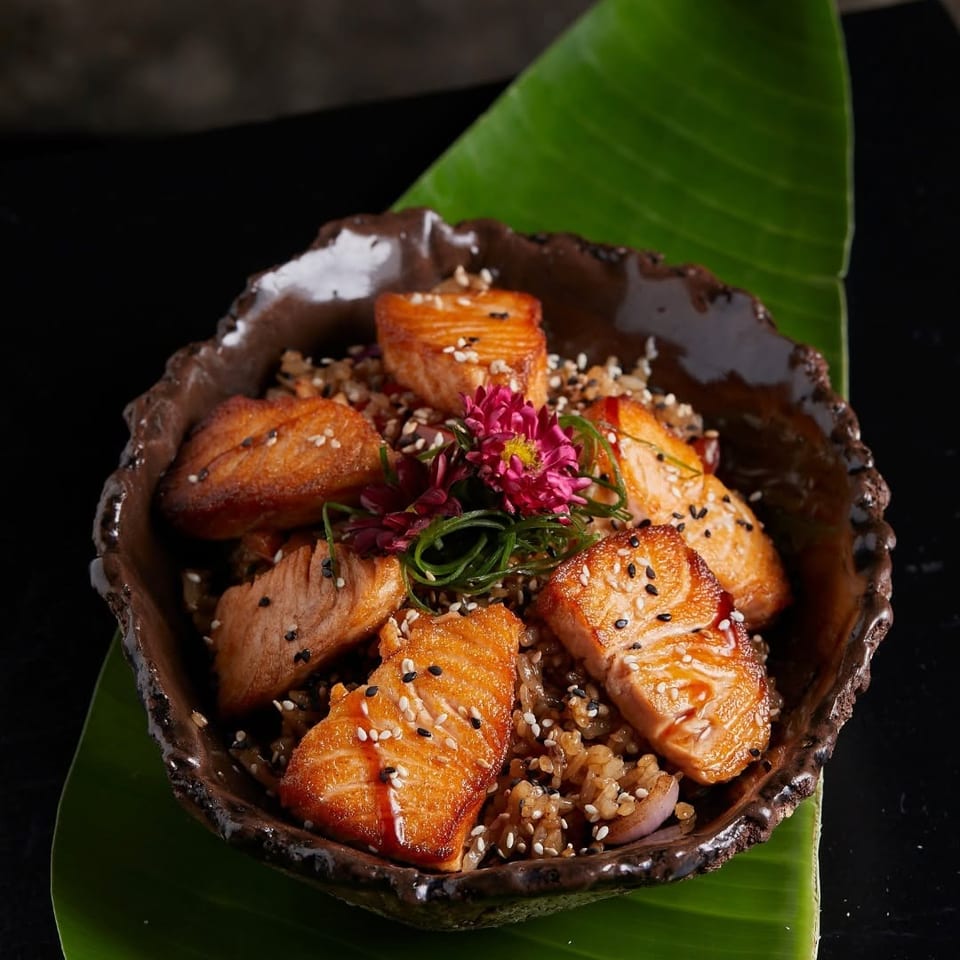 Glazed salmon pieces over seasoned rice, topped with sesame seeds, scallions, and flowers in a rustic bowl on a banana leaf.