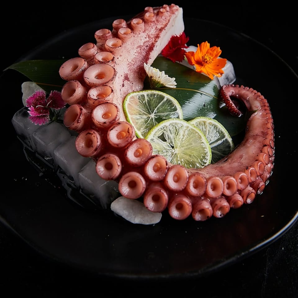 Tentacles of octopus arranged on ice with lime slices, leafy garnish, and colorful flowers on a black plate.